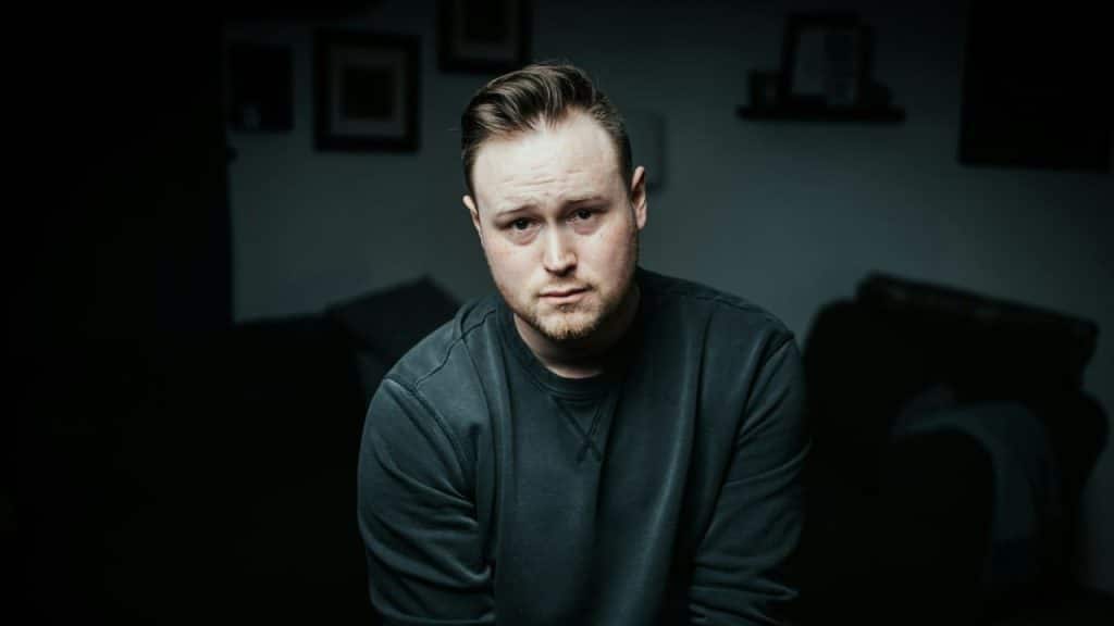 A man with a serious expression looks straight at the camera in a dimly lit room.