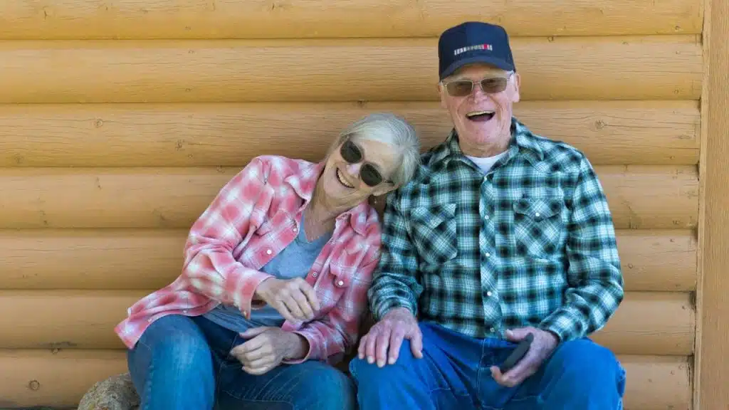 Two older adults in plaid shirts and sunglasses smile while sitting against a log wall.