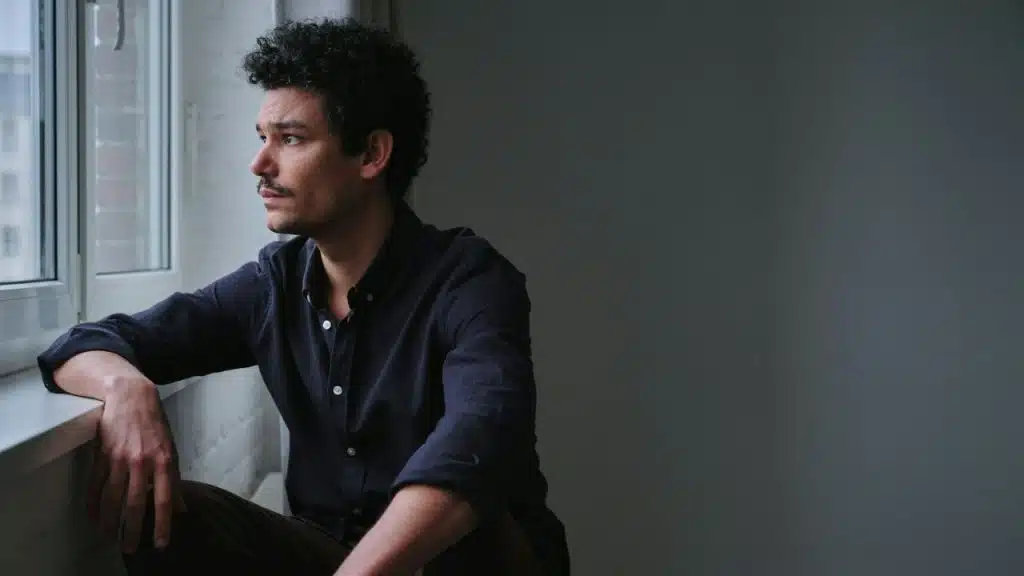 Man with dark curly hair and a mustache sitting by a window, looking outside pensively.