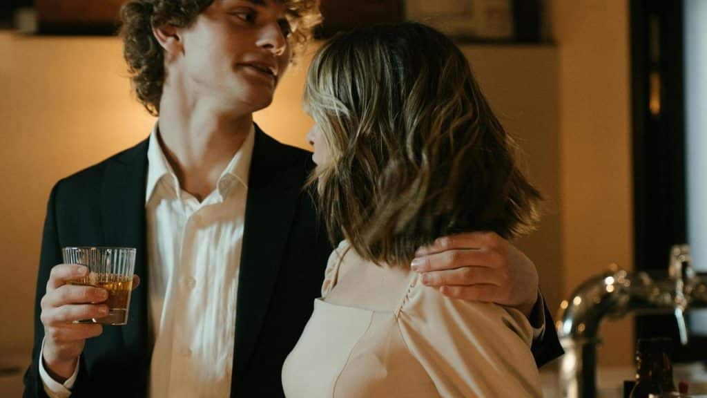 A man in a suit with a drink in hand has his arm around a woman in a light-colored dress.