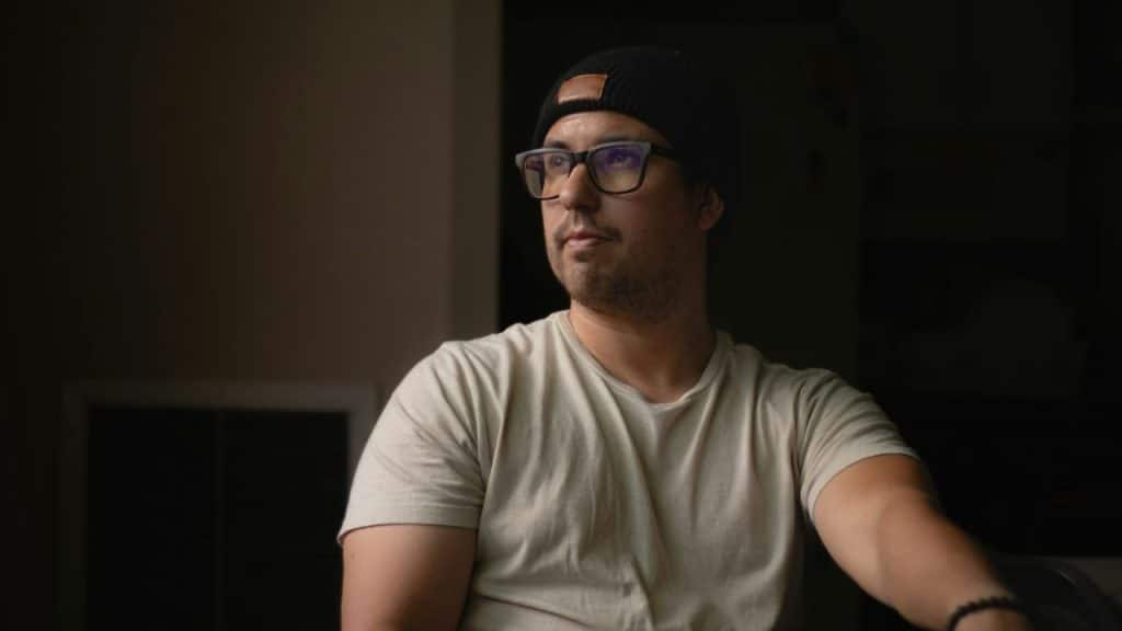 Man in a beanie and glasses looking off-camera in a dimly lit room.