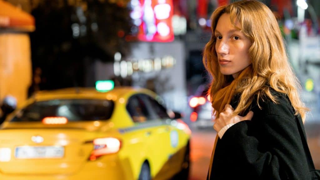 Woman with long wavy hair and a scarf standing on a city street at night next to a yellow taxi.