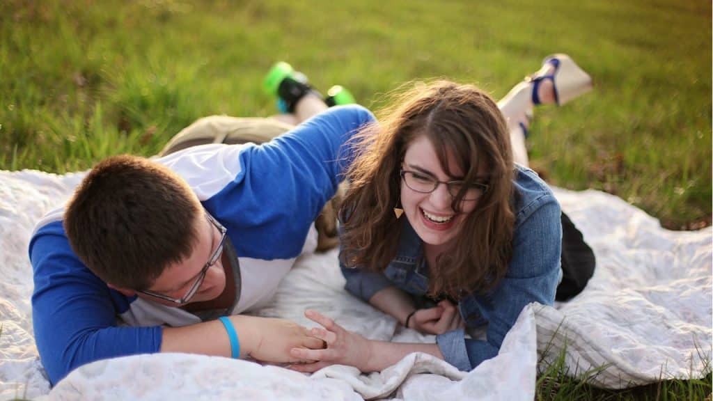 Laughing couple lying on a blanket in the grass holding hands.