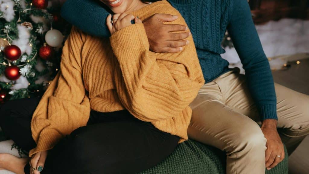 A couple sitting closely together in cozy sweaters beside a decorated Christmas tree.