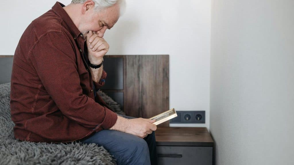 An elderly man sitting on a bed holding a photo frame with a thoughtful expression.