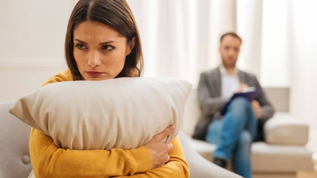 A woman looks resentful while hugging a pillow as a man sits on a couch behind her. .