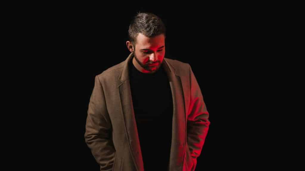 A man in a brown coat and black shirt looks down against a dark background with red light.