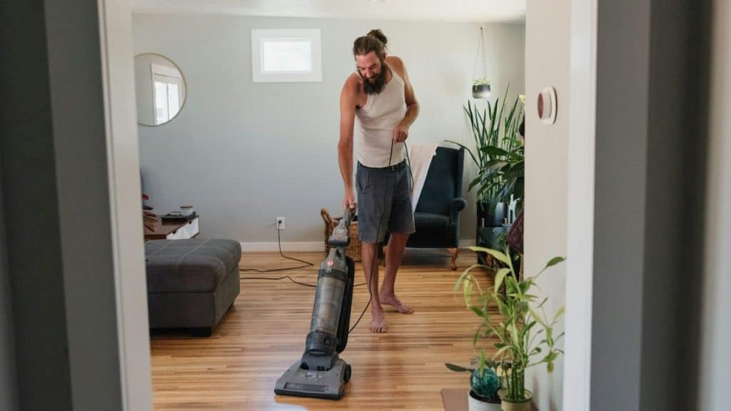 A barefoot man with a beard vacuums a wooden floor in a living room.