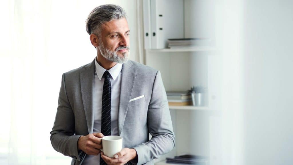 A sharply dressed, bearded man holds a mug and looks thoughtfully out of a bright window.