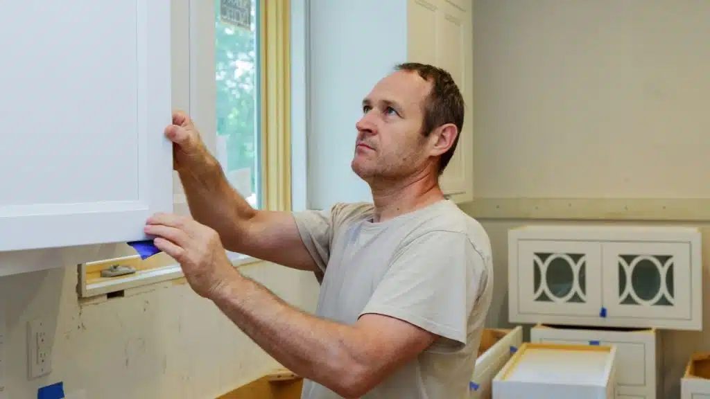 A man installs white kitchen cabinets near a window in a room under construction.