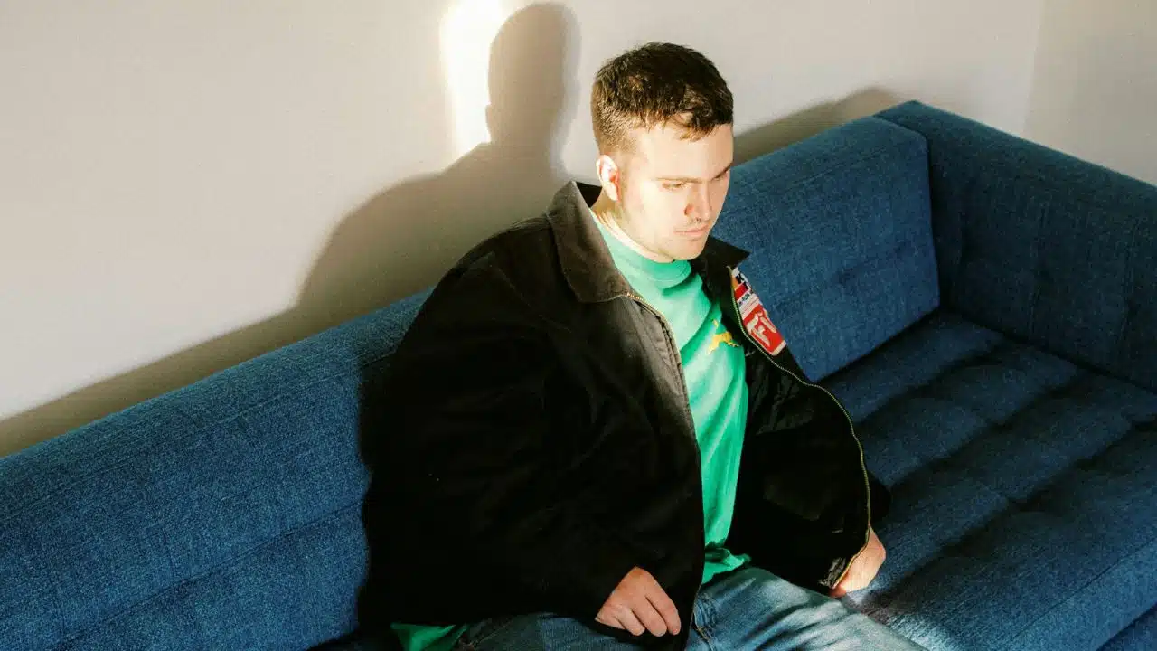 Young man sitting on a blue couch in bright sunlight, wearing a dark jacket over a green shirt.