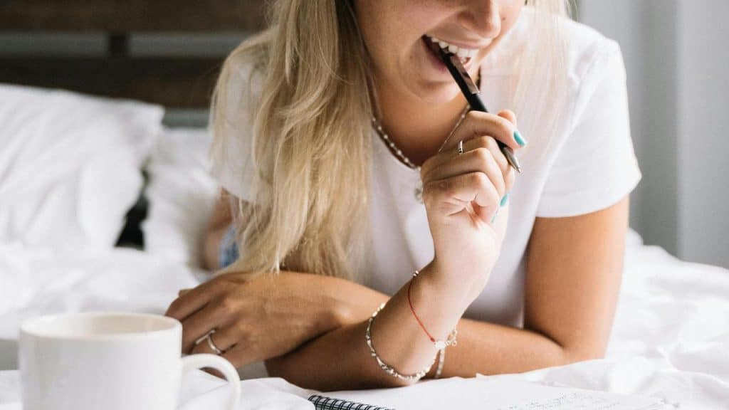 A woman smiling on a bed while holding a pen over a notebook.