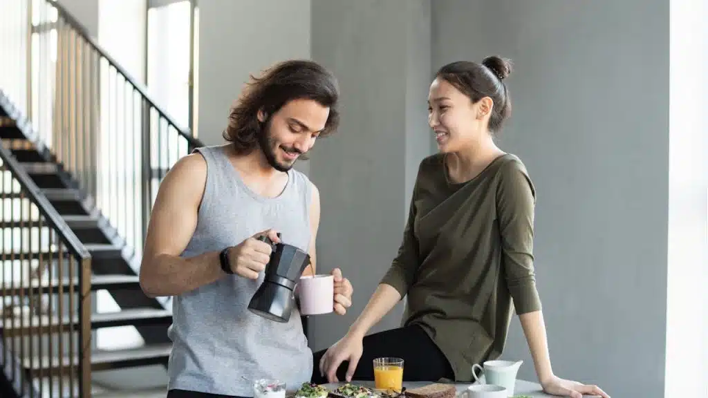 Man pouring coffee for a smiling woman sitting on a kitchen counter near breakfast items.
