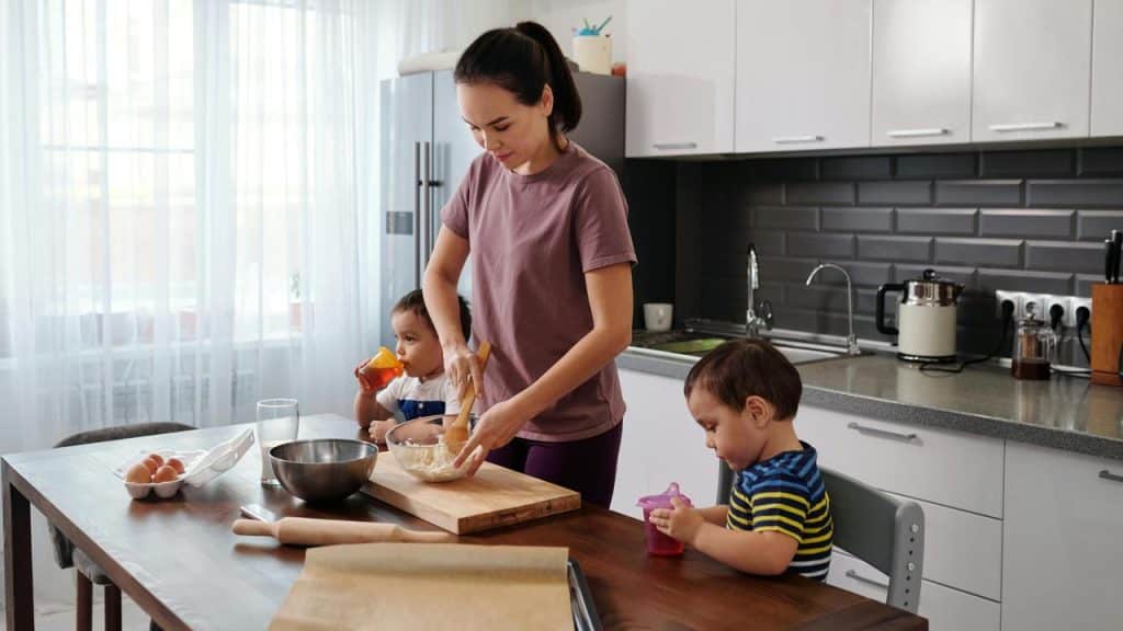 A mother baking in the kitchen while two young children sit and drink nearby.