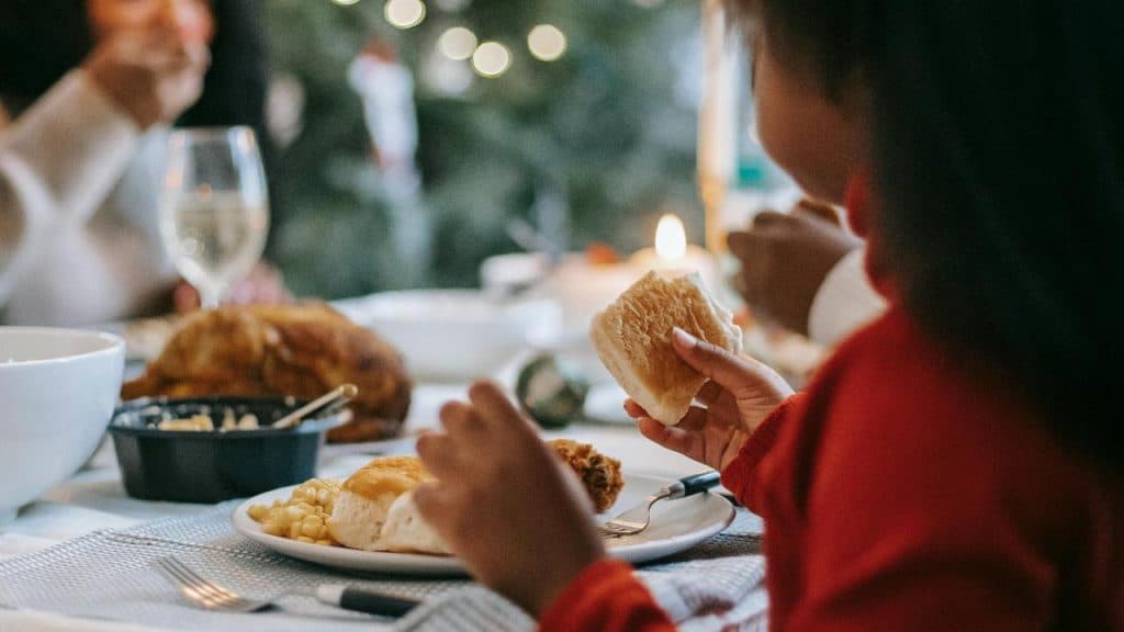 A child eating bread at a holiday dinner table with festive lights in the background.