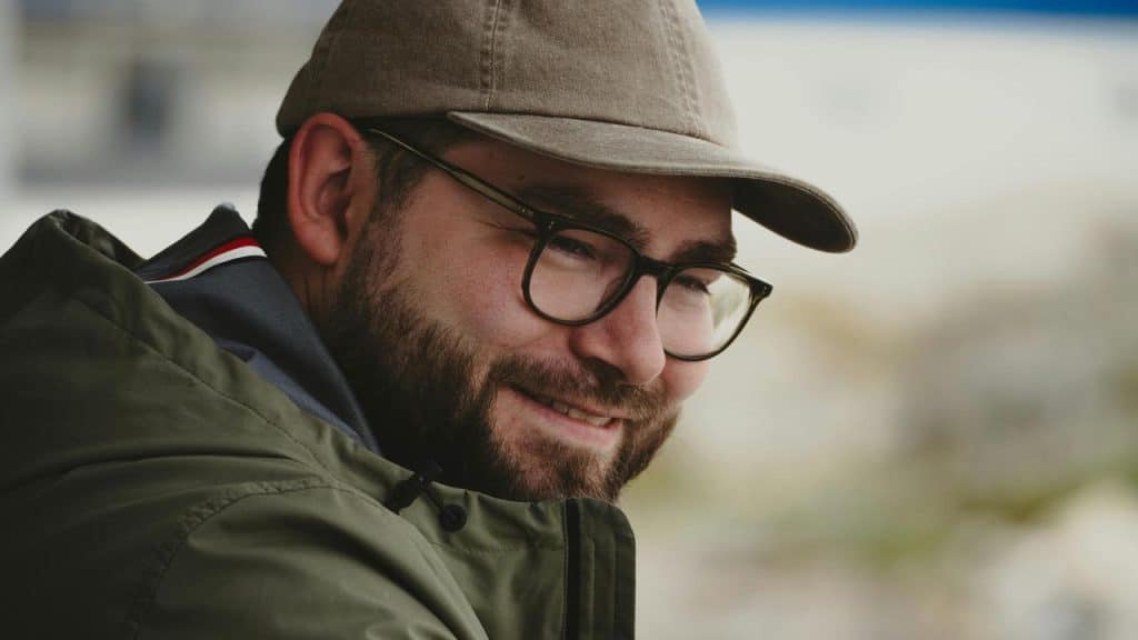 A man in glasses and a cap smiling in a close-up portrait.