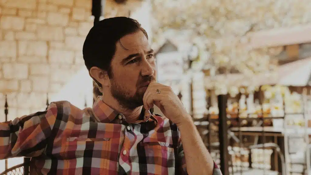 A thoughtful man in a plaid shirt is sitting outdoors, resting his chin on his hand.