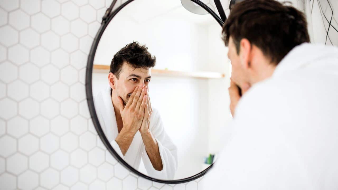 A man in a white robe is washing his face, reflected in a round bathroom mirror.