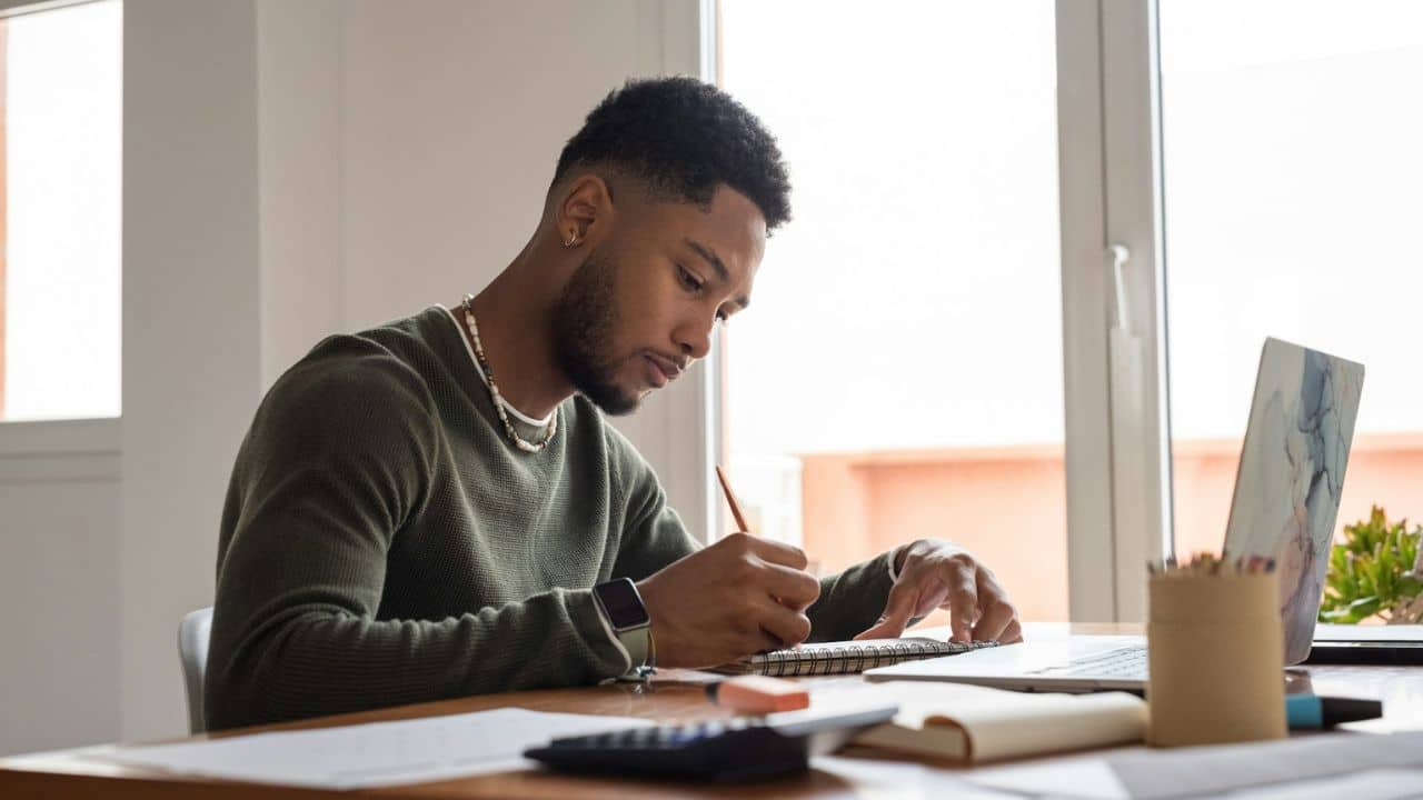 A young man is sitting at a desk by a window, writing in a notebook.