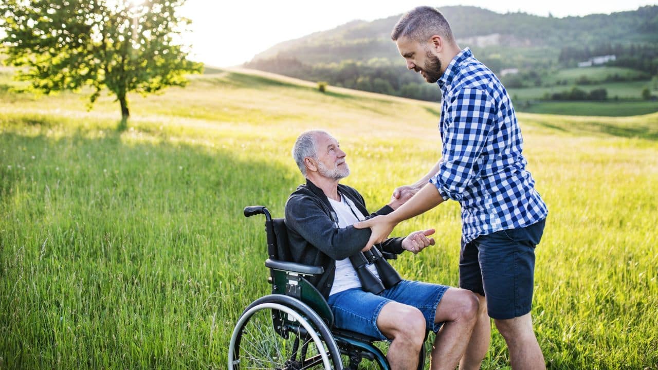 A younger man helping an older man in a wheelchair in a sunny, grassy field.