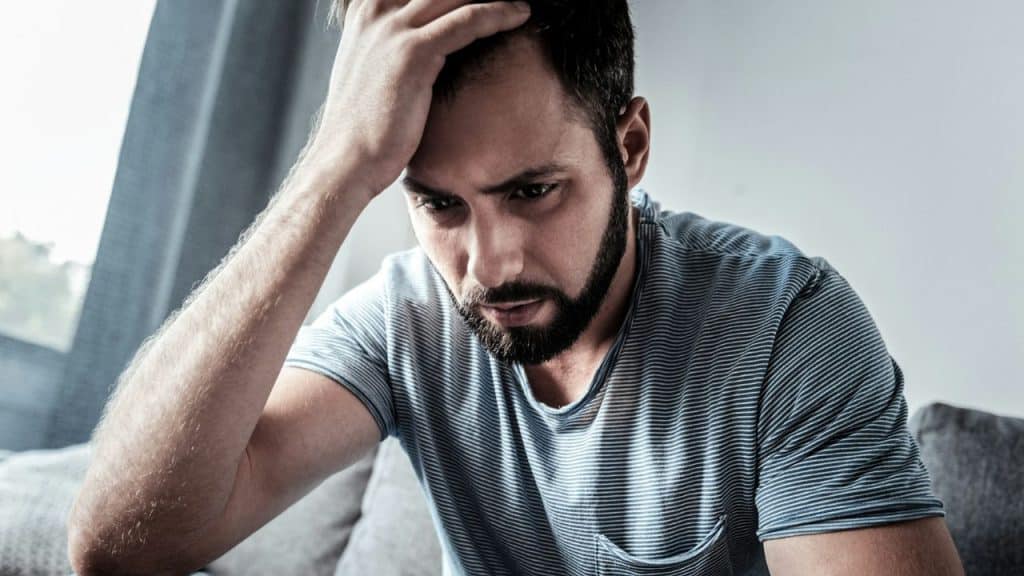 A man with a beard and striped shirt holds his head in apparent distress.