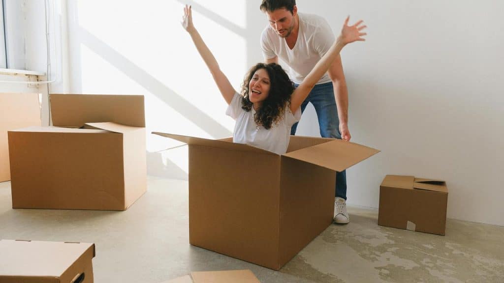 A woman joyfully sitting in a cardboard box as a man stands behind her.