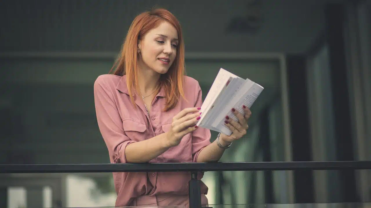 A woman reading a book while standing on a balcony.