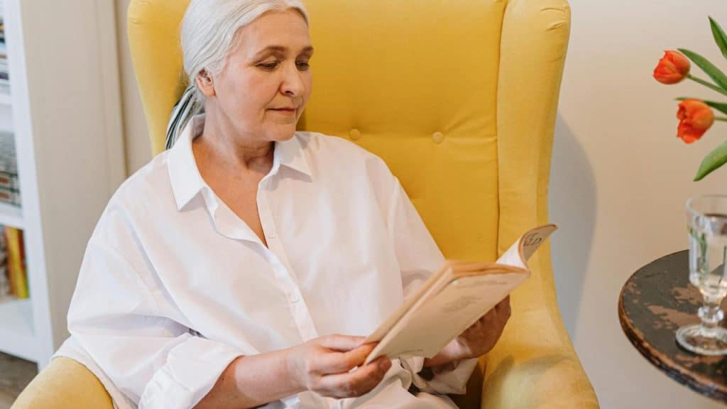 An elderly woman sitting in a yellow chair reading a book.