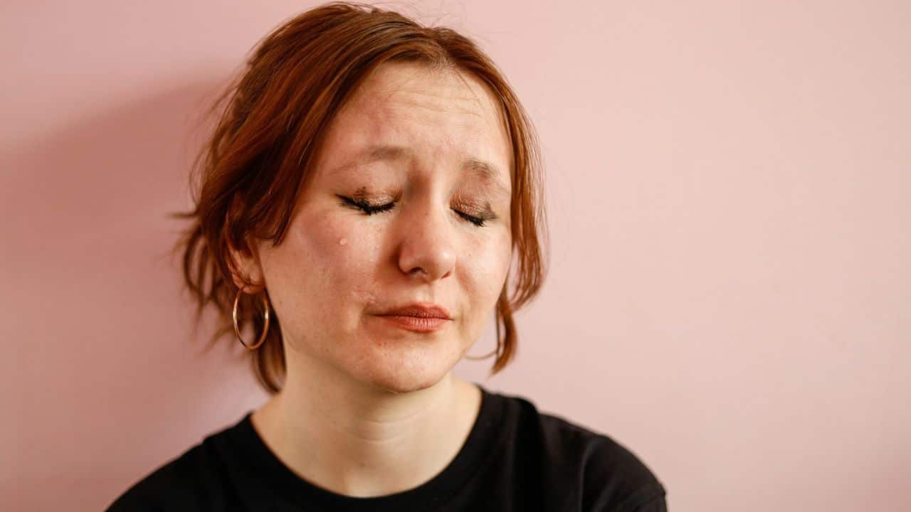 A red-haired person with hoop earrings cries against a pink wall.