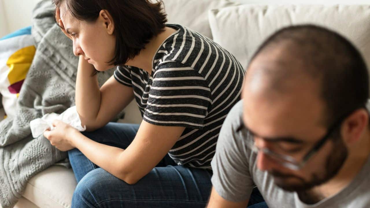A woman sits slumped on a couch holding a tissue near an out-of-focus man.