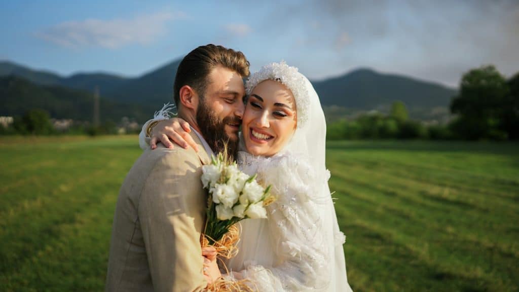 Happy Muslim bridal couple embracing in a sunny green field with mountains in the background.