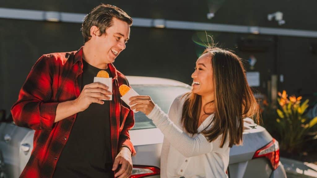 A couple is smiling at each other while standing by a car, holding and eating fried pies or hash browns.