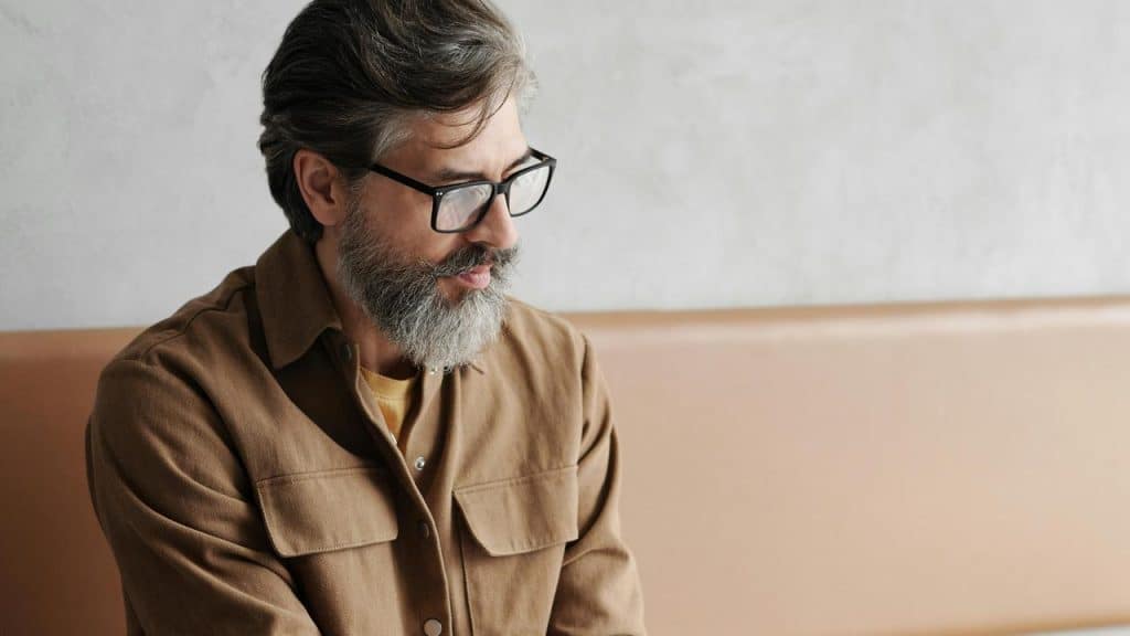 A man with glasses and a beard sitting thoughtfully indoors.
