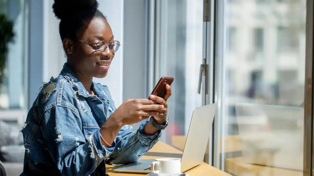 A woman smiling at her phone while sitting by a laptop in a café.