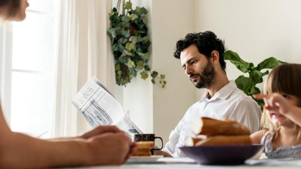 A man reading a newspaper at a breakfast table with his family.