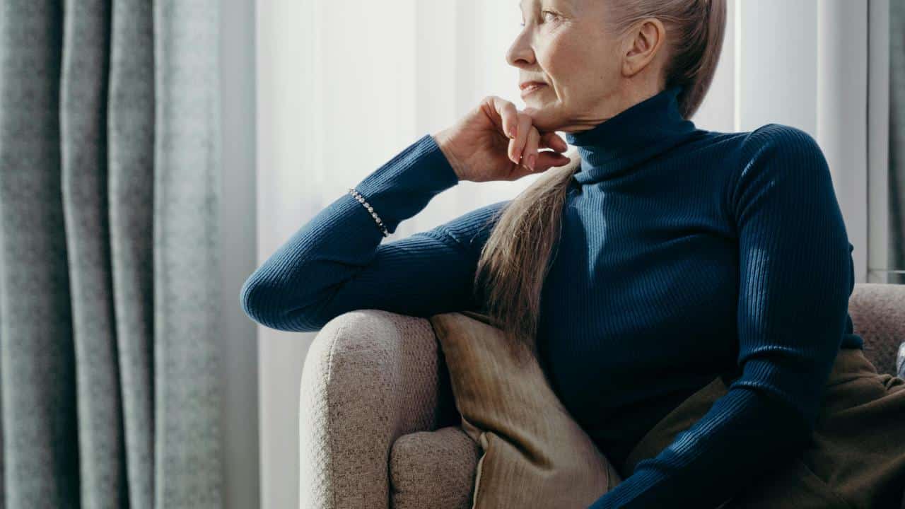 An older woman sitting on a couch and gazing thoughtfully to the side.