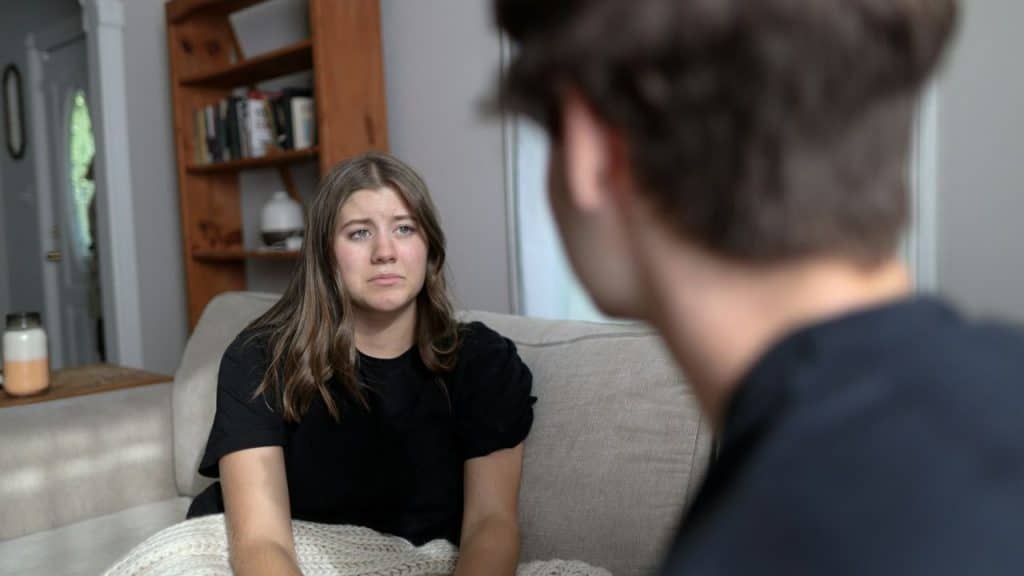 A distressed woman sits on a couch, looking at a person whose back is in the foreground.
