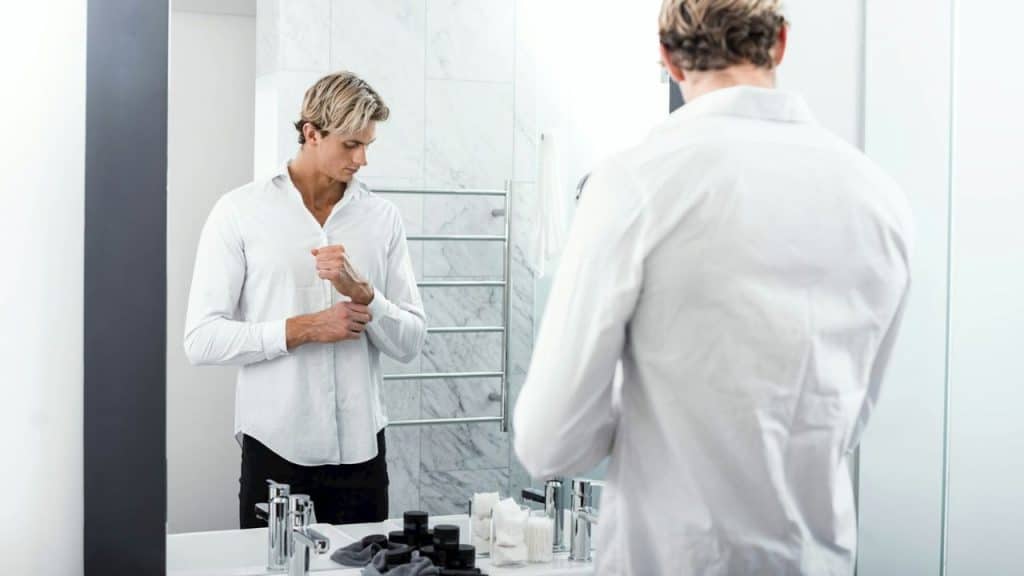 A man with blonde hair buttons his white shirt while looking in a bathroom mirror.