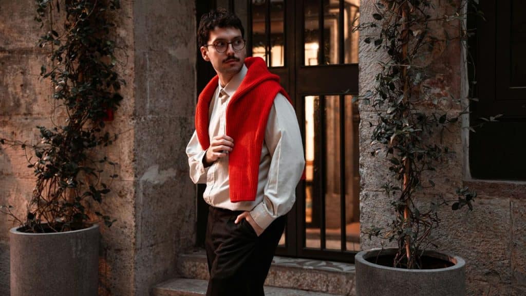 Man with glasses and mustache stands outside with a red sweater draped over his shoulders.