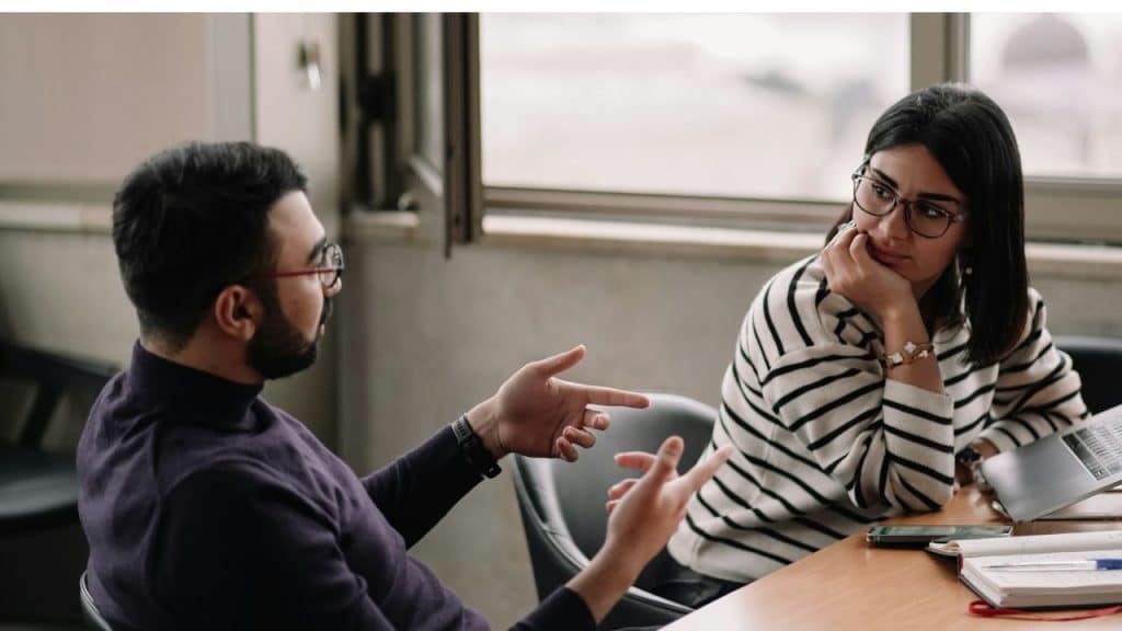 A man gestures while talking to a woman wearing glasses, who rests her chin on her hand.