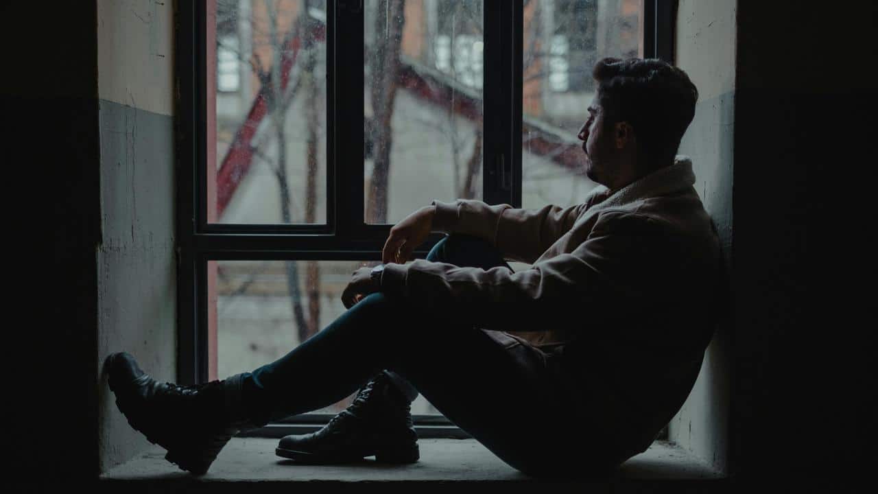 A man sitting in a dark windowsill, gazing outside thoughtfully.
