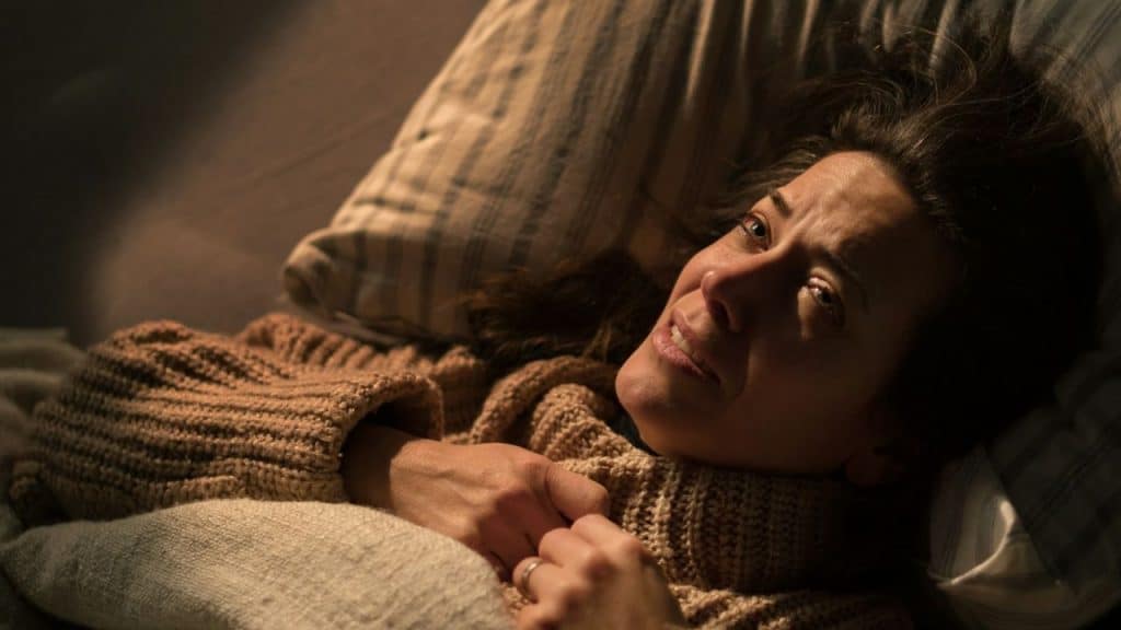 Woman in a brown sweater lying in bed looking up with a distressed expression.