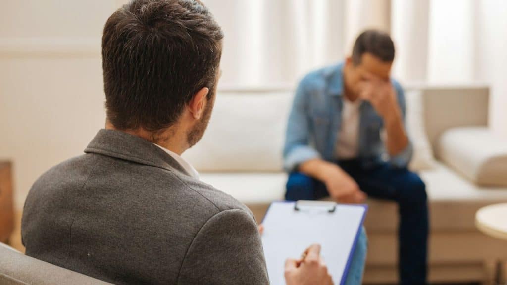 Therapist holding a clipboard facing a distressed man sitting on a sofa.