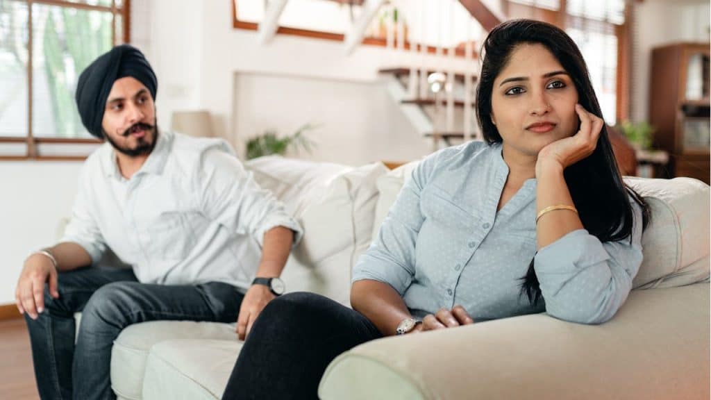 A woman sits on a couch while a man in a turban sits nearby looking at her with a concerned expression.