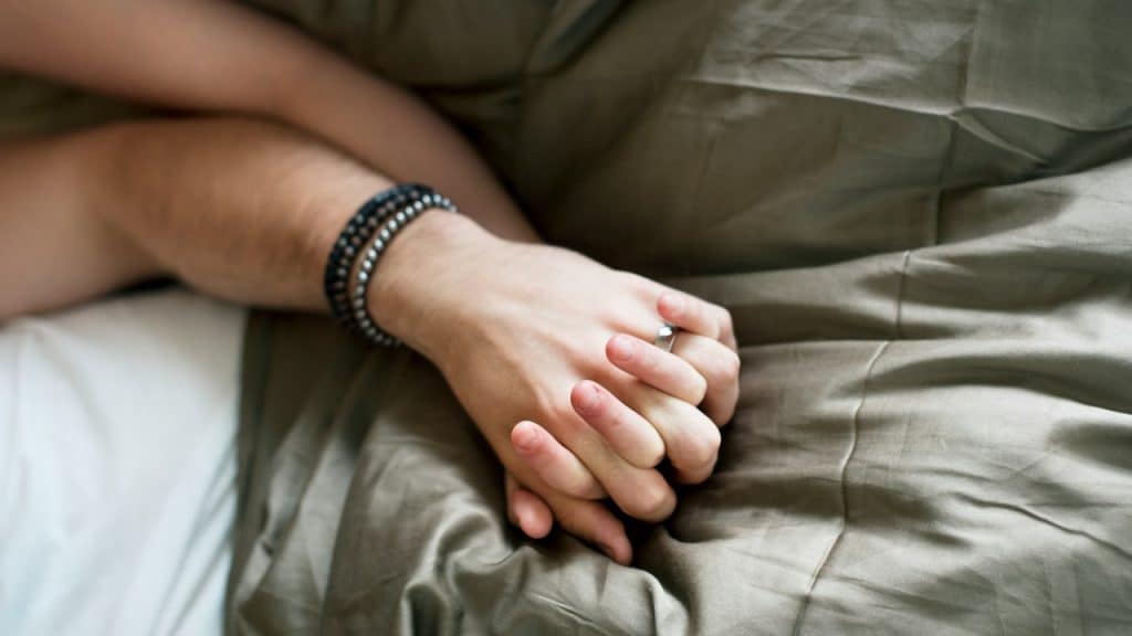 A close-up shot of two people holding hands while lying in bed under a duvet.