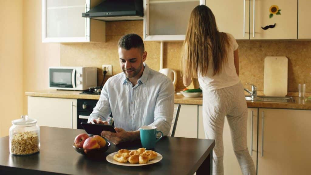Man smiling while using a tablet at a table as a woman works in the kitchen.