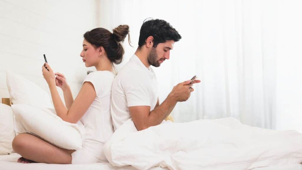 A man and woman are sitting on a bed back to back, each using a mobile phone.