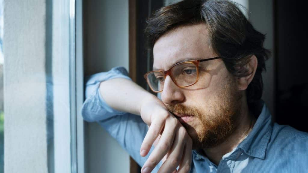 A man with glasses and a beard looks thoughtfully out a window.