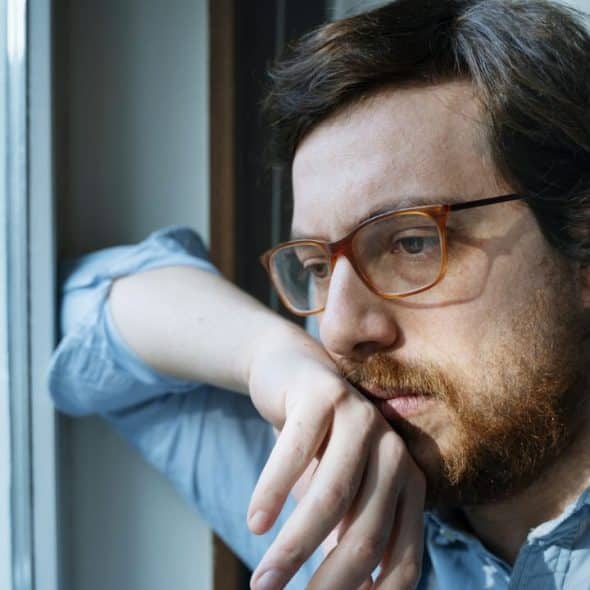 A man with glasses and a beard looks thoughtfully out a window.