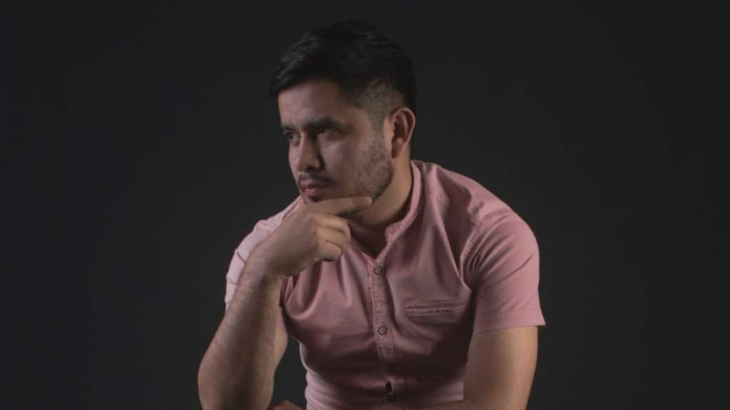 Man in a pink shirt looking away with his hand resting on his chin.
